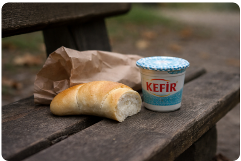 Egyszerű kifli és rendszerváltás kori kefír egy parkpadon, papírzacskó mellett.